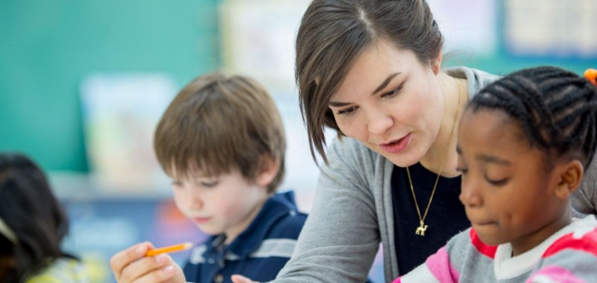 Lehrerin erklärt einer Schülerin etwas, im Hintergrund liest ein anderer Schüler etwas für sich