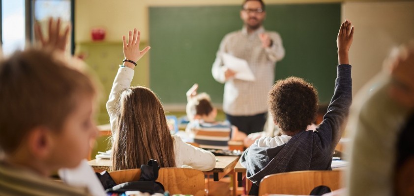 Ein Lehrer steht vor einer Klasse von jungen Kindern an der Tafel und fragt etwas ab. Mehrere Kinder melden sich. 