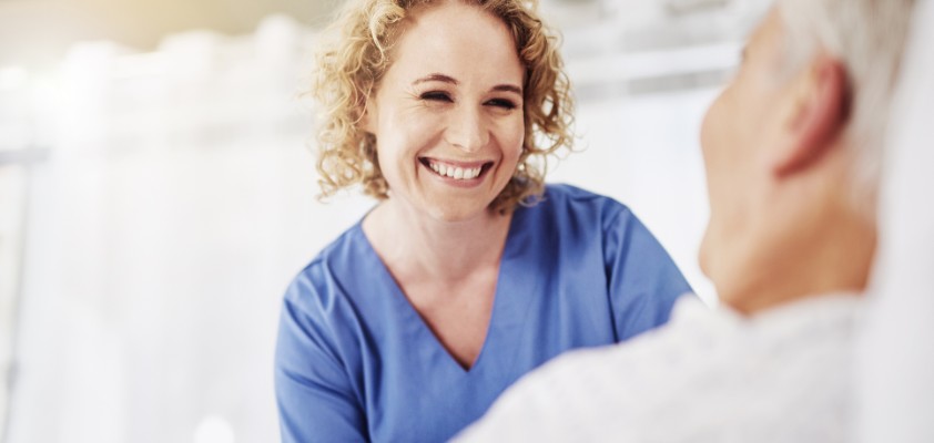 Eine Pflegerin schaut lächelnd einen im Bett liegenden Patienten an. 