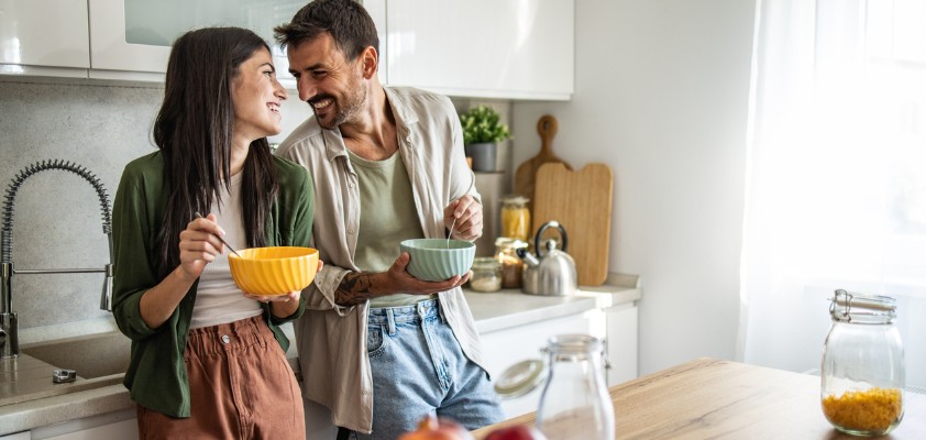 Ein junges Paar steht mit Müslischalen in der Hand steht lachend in der Küche
