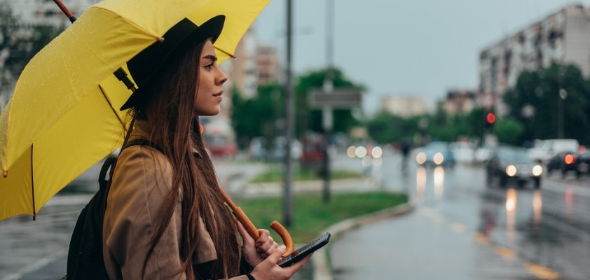 Eine junge Frau steht im Regen mit einem gelben Regenschirm am Straßenrand und sucht mit ihrem Handy in der Hand etwas. 