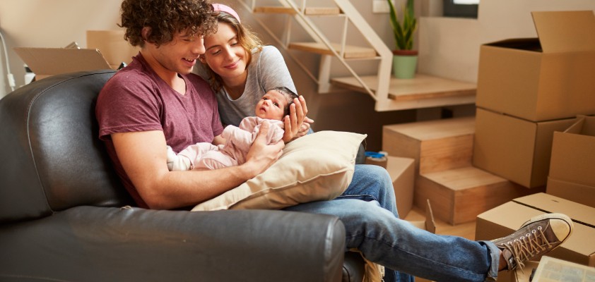 Junges Pärchen mit Baby im Arm sitzen im Wohnzimmer, voll mit Umzugskartons