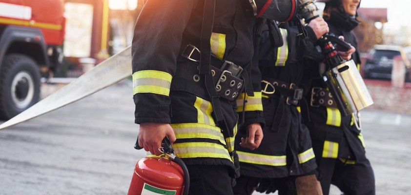 Drei Feuerwehrleute laufen mit Equipment herum. Ihre Uniformen und Equipment stehen im Fokus des Bildes. 