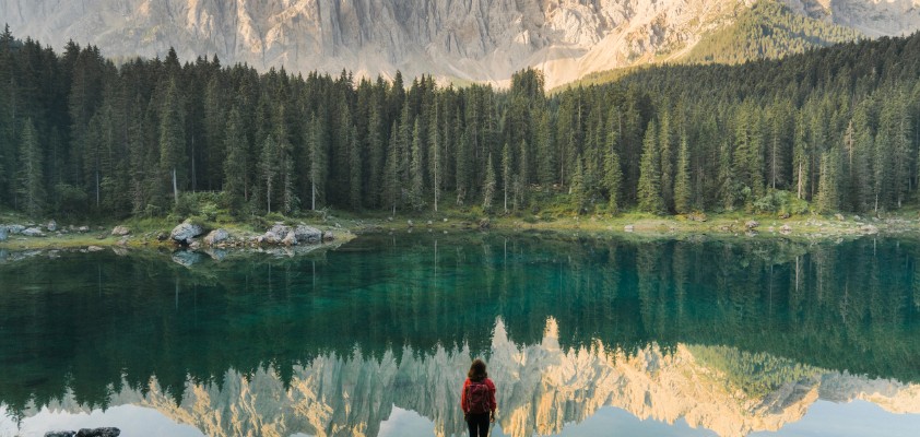 Person in roter Jacke steht am Ufer eines Sees, der von Bergen und Wäldern umgeben ist.