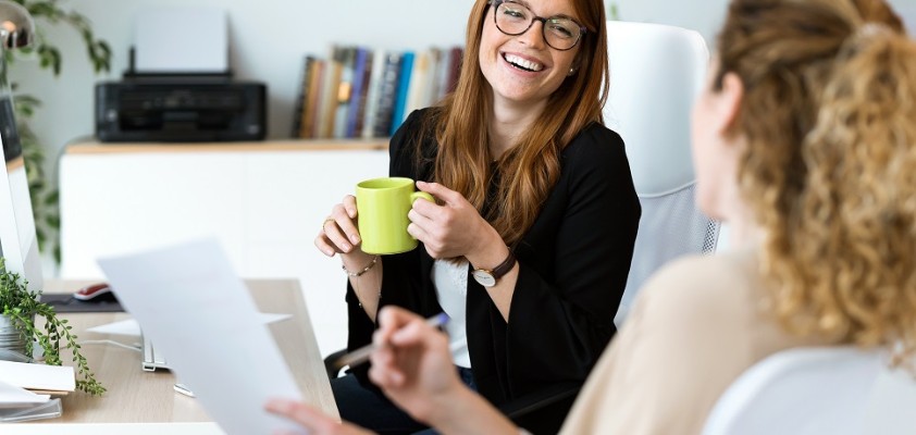 Zwei junge Frauen unterhalten sich lachend am Arbeitsplatz.