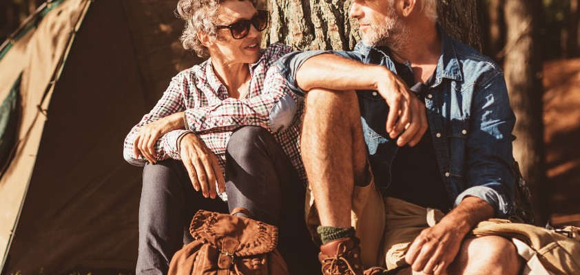 Eine Frau ein Mann in Wanderkleidung sitzen nebeneinander angelehnt an einen Baum und unterhalten sich.