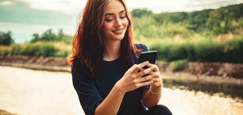 Eine junge Frau schaut am Ufer eines kleinen Flusses auf ihr Smartphone.