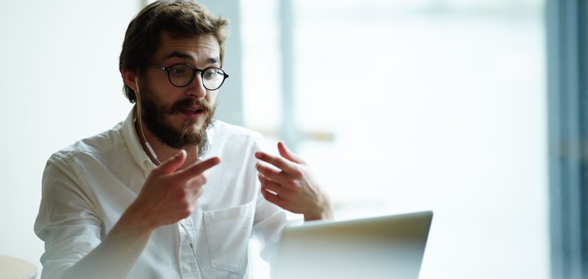 Ein Mann sitzt am Laptop in einem Online-Meeting und benutzt seine Hände für Gestiken. 