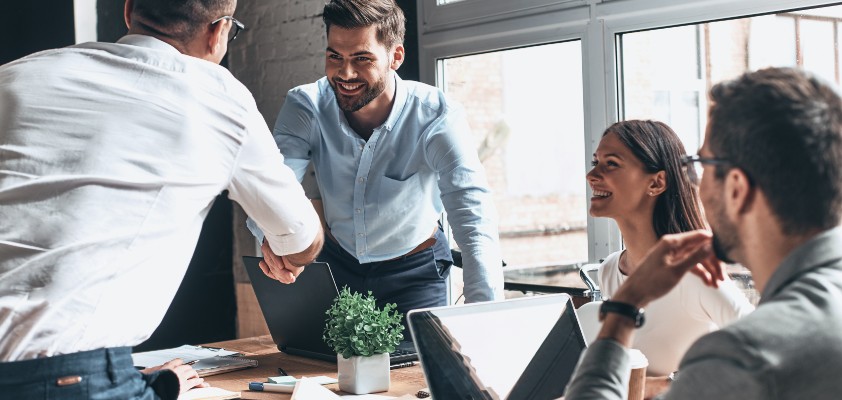Eine kleinere Gruppe von Leuten sitzen zusammen im Büro und zwei Personen reichen sich die Hände. Sie tragen alle Business-Kleidung. 