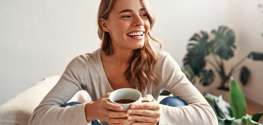 Eine Frau sitz lächelnd mit einer Tasse Kaffee auf dem Sofa.