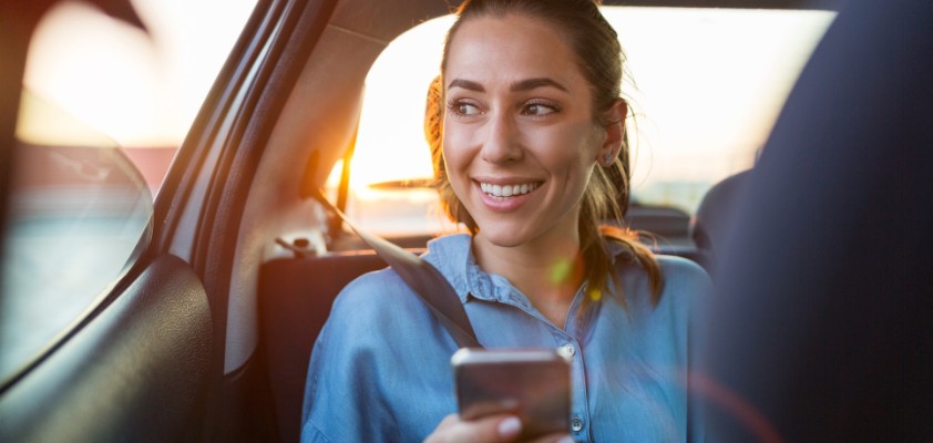 Frau sitzt auf der Rückbank, schaut lachend aus dem Fenster hinaus während sie ein Telefon in der Hand hält.