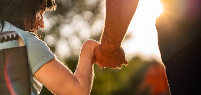Person im Rollstuhl hält die Hand einer anderen Person im Sonnenlicht.