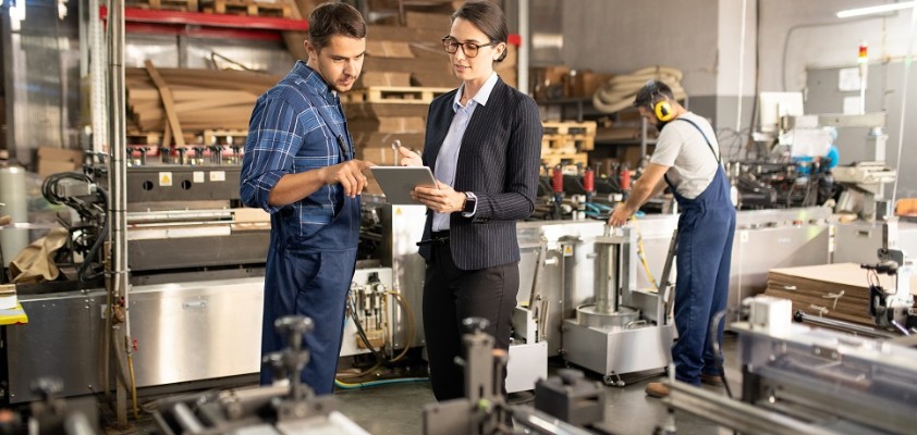 Eine Business-Frau und ein Bauarbeiter sprechen zusammen über ein Dokument, welches die Frau in der Hand hält. Die Umgebung ist eine Fabrik. 