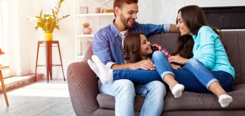 Zwei junge Eltern sitzen lachend mit ihrer Tochter auf dem Sofa.