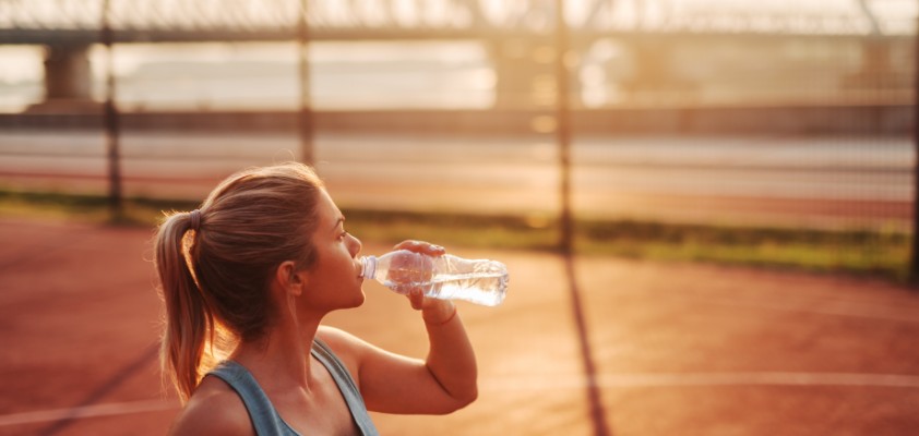 Eine junge Frau steht in einem kurzen Top auf einem Basketballfeld und trinkt Wasser aus einer Flasche.