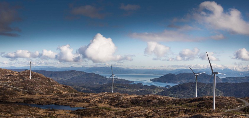 Landschaft mit Windkraftanlagen auf Hügeln und Blick auf das Meer im Hintergrund.