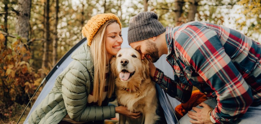 Frau mit gelber Mütze und Mann mit kariertem Hemd beim Camping mit Golden Retriever