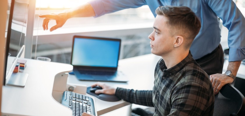 Ein junger Mann sitzt am PC und ein älterer Mann zeigt ihm etwas. 