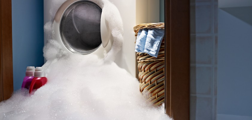 Bühnenbild Ratgeber Kuriose Missgeschicke  Das Bild zeigt eine überlaufende Waschmaschine, die mit Schaum bedeckt und umgeben ist.
