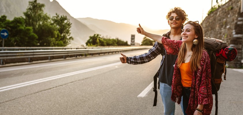 Zwei Backpacker stehen mit ihren Rucksäcken am Straßenrand und halten ihren Daum raus. Sie warten auf eine Mitfahrgelegenheit. 
