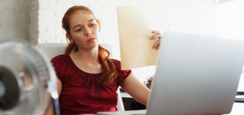 Eine Frau sitzt verschwitzt am Laptop, vor ihr ein Ventilator und sie fächert sich Luft mit Unterlagen zu. 