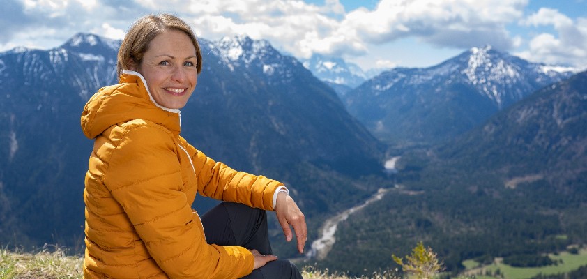 Magdalena Neuner sitzt auf einer Wiese in den Bergen und blickt in die Kamera.