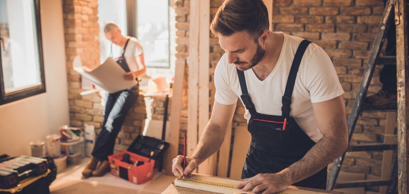 Ein Mann arbeitet auf einer Baustelle und misst ein Stück Holz ab. 