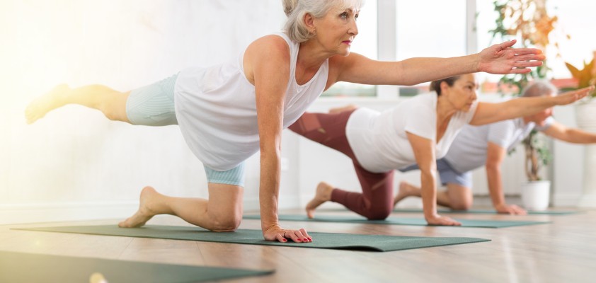 Ältere Frau macht mit anderen eine Balance-Übung auf Yogamatten in einem hellen Raum.
