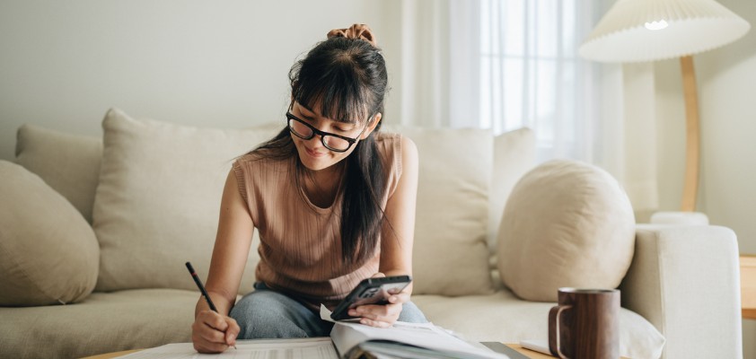 Eine junge Frau sitzt auf einem Sofa, schaut auf Unterlagen auf dem Tisch und macht Notizen, während sie ein Smartphone in der Hand hält.