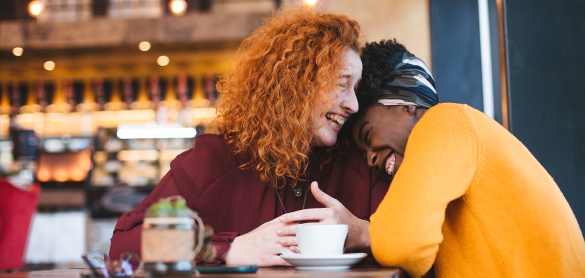 Zwei Frauen sitzen in einem Café und liegen sich lachend in den Armen. 
