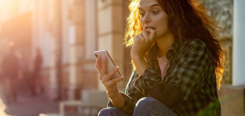 Eine junge Frau sitzt draußen auf einer Steinstufe und schaut gespannt in ihr Smartphone. 