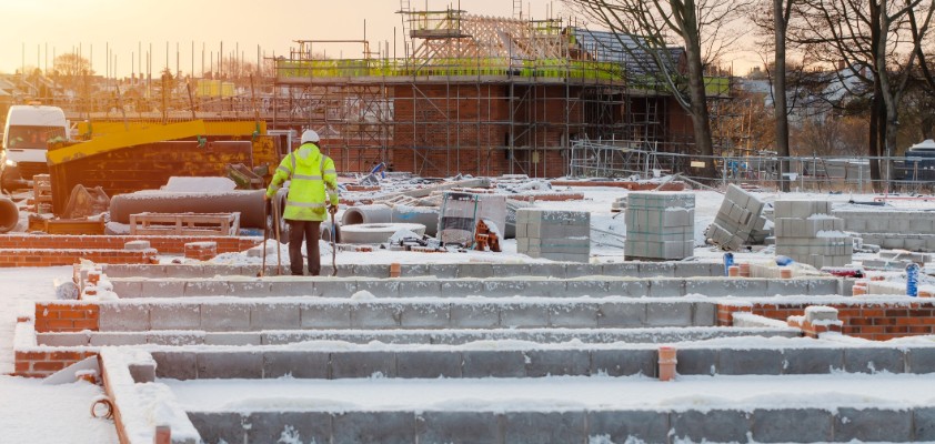 Dach einer verschneiten Baustelle. In Ferne kann ein Bauarbeiter in gelber Warn-Schutzkleidung erkannt werden. 