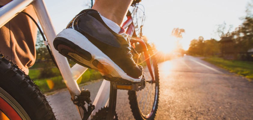 Ein Mann fährt sein Fahrrad im Sonnenuntergang auf einem asphaltierten Weg. Im Fokus stehen seine Sportschuhe.
