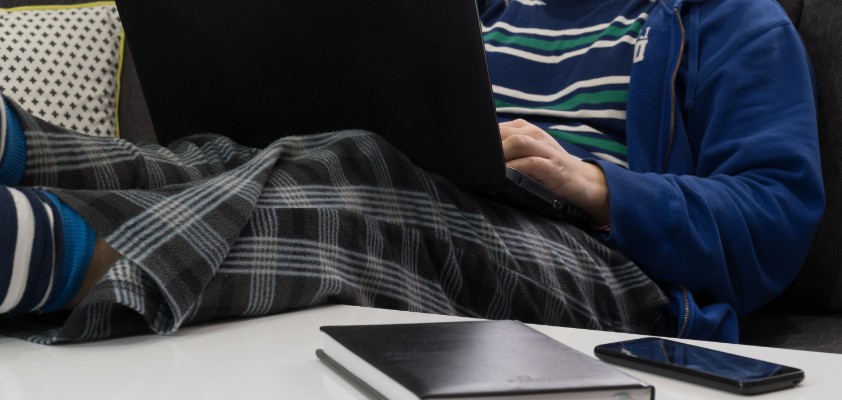 Eine männliche Person (Gesicht nicht zu sehen) liegt auf dem Sofa in Jogginghose und arbeitet am Laptop.