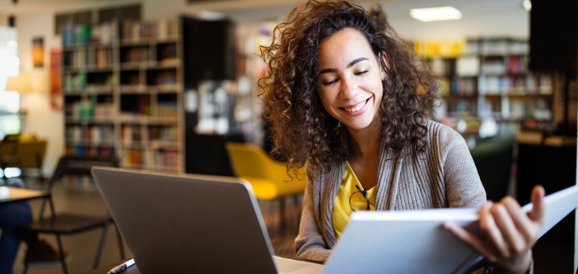 Eine junge Frau sitzt in einem Buchladen am Laptop und schaut währenddessen in ein Buch. 