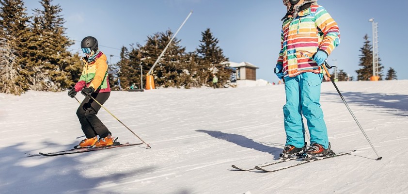 Zwei Kinder, die nebeneinander her auf einer Skipiste Ski fahren. 