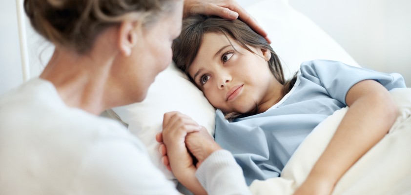 Ein kleiner Junge liegt im Krankenbett. Seine Mutter sitzt daneben und streichelt ihm den Kopf.