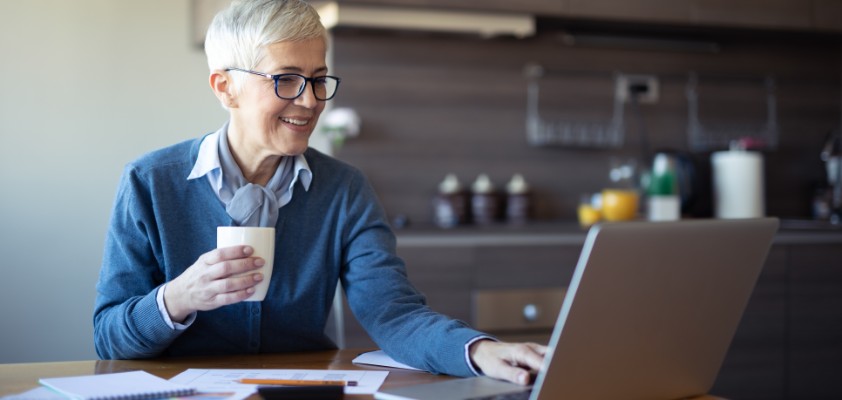 Eine ältere Frau sitzt lächelnd mit einem Kaffee in der Hand am Laptop.