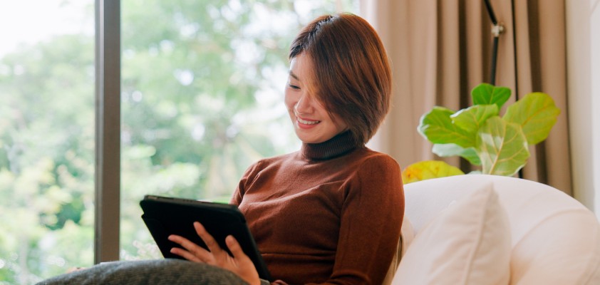 Eine Frau sitzt entspannt auf dem Sofa und schaut auf ihr Tablet