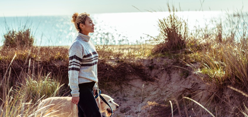 Eine Frau geht mit einem Hund an der Küste entlang, umgeben von Dünen und Meer im Hintergrund.