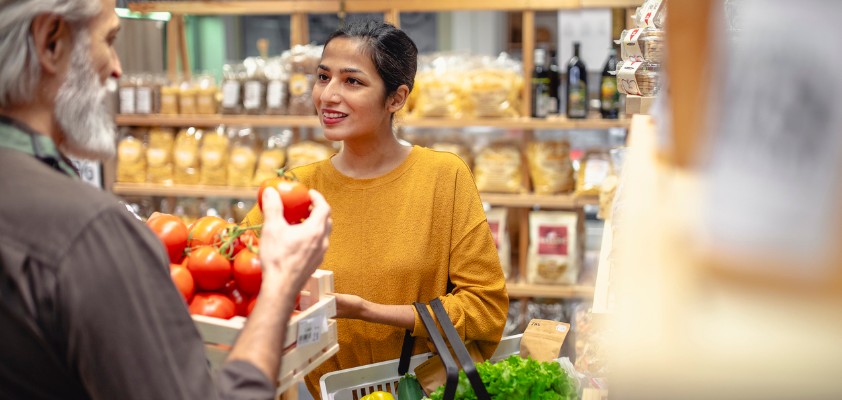 Eine junge Frau lächelt, während sie mit einem älteren Mann in einem Laden spricht und dabei einen Korb mit frischen Lebensmitteln hält. Im Hintergrund sind Regale mit verpackten Produkten zu sehen.