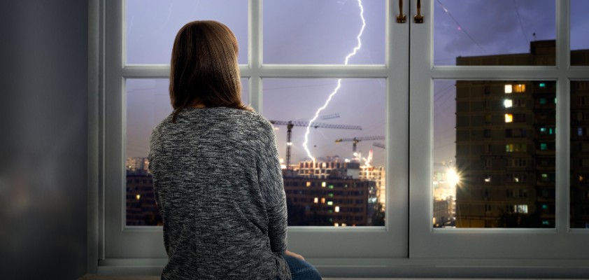 Ein Mädchen sitzt knieend an einem Fenster und schaut die sichtbaren Blitze in der Ferne an. 
