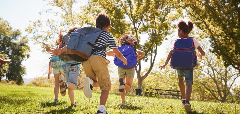 Mehrere Kinder laufen mit ihren Schulrucksäcken über eine Wiese in der Sonne.