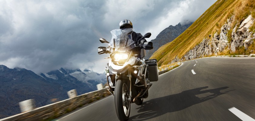 Motorradfahrer fährt auf einer kurvigen Bergstraße, im Hintergrund sind Berge und dunkle Wolken zu sehen.