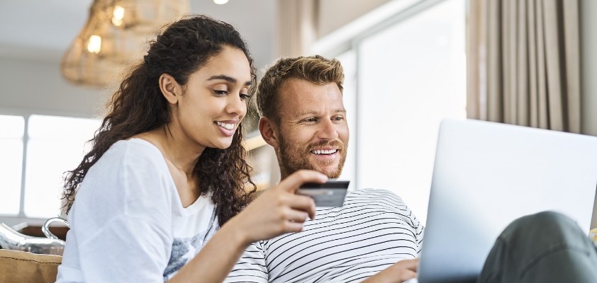 Eine Frau und ein Mann sitzen zusammen auf einem Sofa. Der Mann hält einen Laptop auf seinem Schoß und die Frau hält eine Kreditkarte in der Hand. 