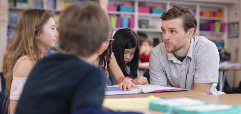 Ein junger Lehrer unterhält sich  mit zwei Schülern. Sie sitzen zu dritt an einem Tisch in einer Bibliothek. 