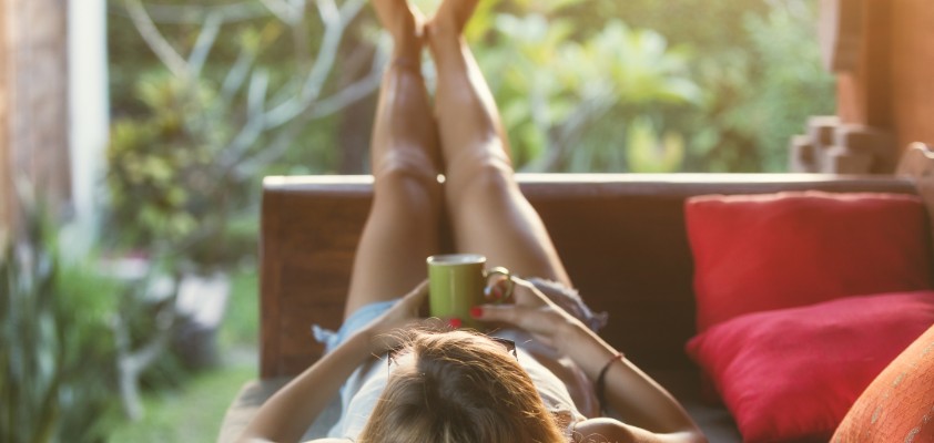 Eine Frau liegt entspannt auf einem Sofa auf einer Veranda und legt die Beine hoch.