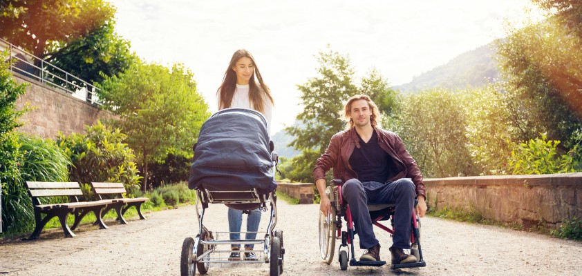 Eine Frau schiebt einen Kinderwagen, während ein Mann daneben in einem Rollstuhl sitzt. Sie spazieren gemeinsam einen Weg entlang. 