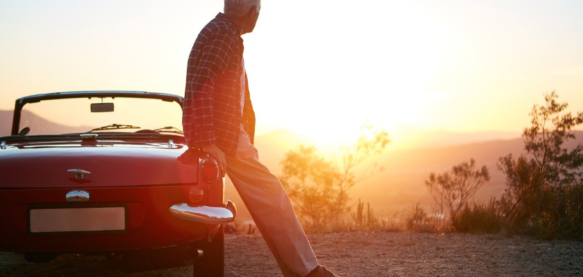 Ein erwachsener Mann lehnt an seinem Oldtimer und schaut von einem Hügel aus in den Sonnenuntergang. 