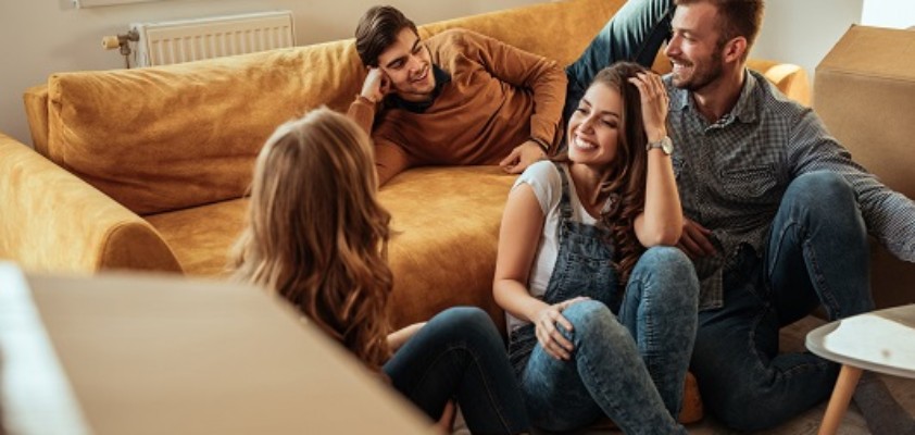 Eine Gruppe von vier Freunden sitzen zusammen zwischen Umzugskartons in einem leeren Wohnzimmer.